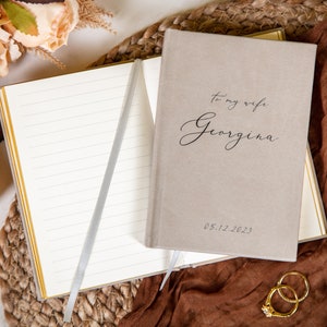 May include: A beige notebook with the inscription "To my wife Georgina" and the date 05.12.2023. The notebook is open, revealing lined pages with a silver ribbon bookmark. Gold pen, rings, and flowers are arranged around the notebook.