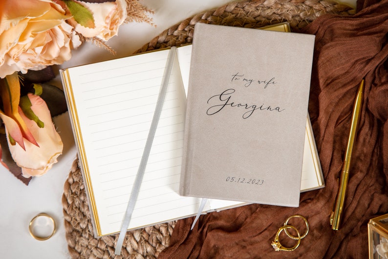 May include: A beige notebook with the inscription "To my wife Georgina" and the date 05.12.2023. The notebook is open, revealing lined pages with a silver ribbon bookmark. Gold pen, rings, and flowers are arranged around the notebook.