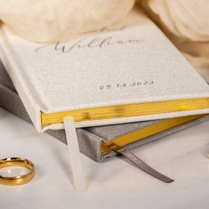 May include: A stack of two hardcover journals with gold gilded edges. The top journal is off-white with the word "Willkommen" and the date "05.12.2023" printed on the cover. A gold ring and pen are also visible.