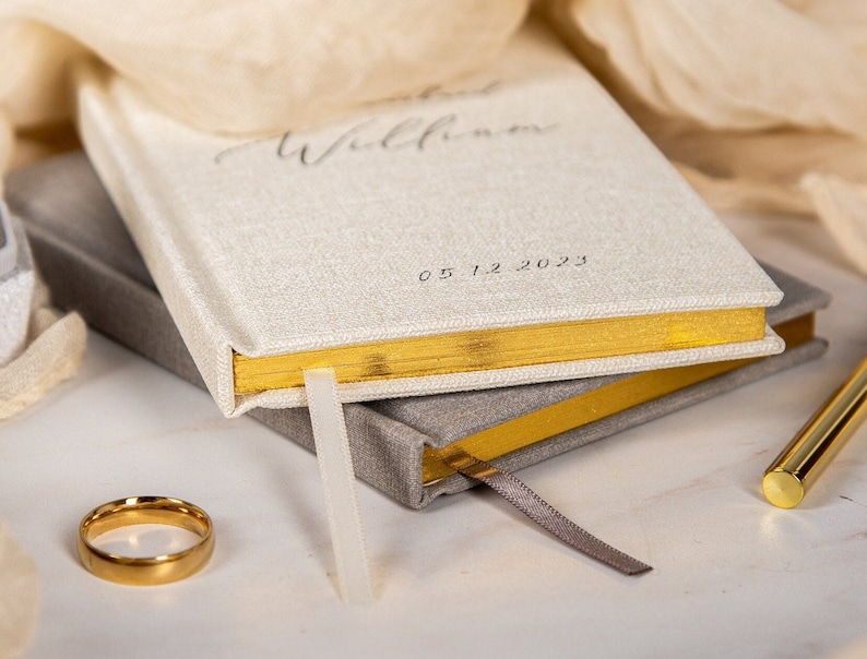 May include: A stack of two hardcover journals with gold gilded edges. The top journal is off-white with the word "Willkommen" and the date "05.12.2023" printed on the cover. A gold ring and pen are also visible.
