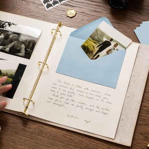 May include: An open scrapbook with gold-colored rings, holding black and white photos and a blue envelope with a photo inside. A handwritten note is visible on the page. A gold pen and other stationery items are on the wooden surface.