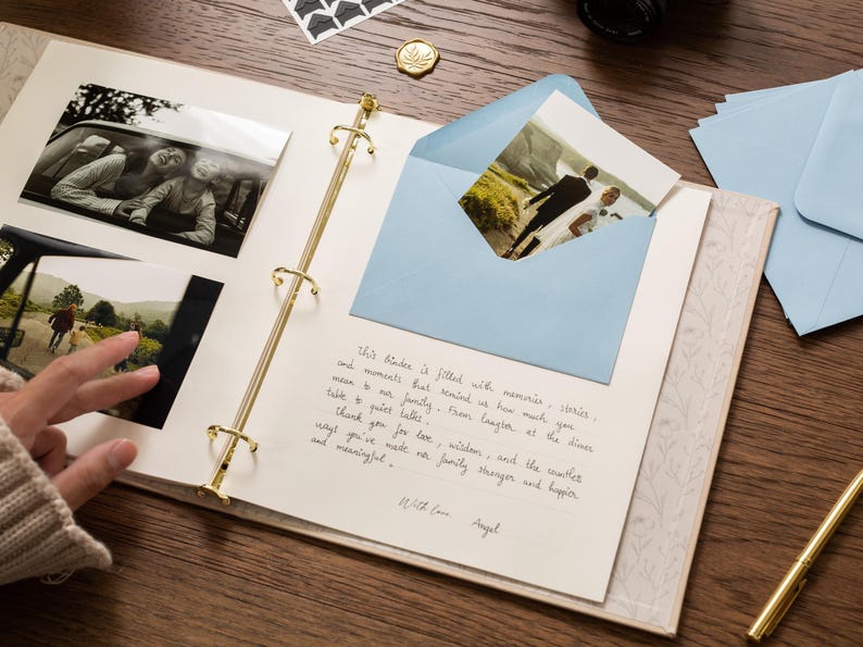 May include: An open scrapbook with gold-colored rings, holding black and white photos and a blue envelope with a photo inside. A handwritten note is visible on the page. A gold pen and other stationery items are on the wooden surface.