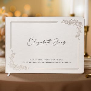 May include: Cream-colored memorial guest book with the name "Elizabeth Jones" in elegant script. The dates "May 13, 1979 - November 14, 2022" are below the name, along with the words "Loved beyond words, missed beyond measure."