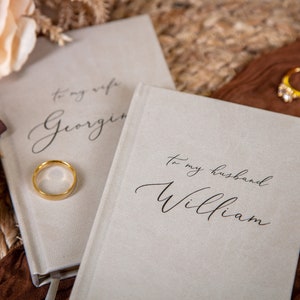 May include: Two light gray notebooks with the inscriptions "To my wife Georgina" and "To my husband William" in elegant script. A gold wedding band rests on the first notebook, with two more rings visible in the background. A gold pen is also present.