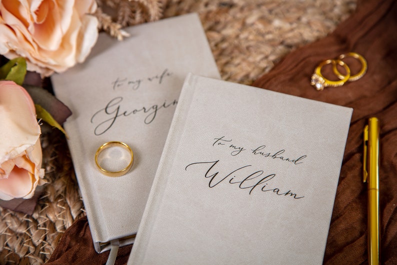 May include: Two light gray notebooks with the inscriptions "To my wife Georgina" and "To my husband William" in elegant script. A gold wedding band rests on the first notebook, with two more rings visible in the background. A gold pen is also present.
