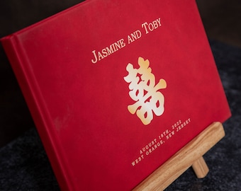May include: A red wedding album with gold lettering that reads "Jasmine and Toby" and a gold double happiness symbol. The album also includes the date August 18th, 2022, and the location West Orange, New Jersey.