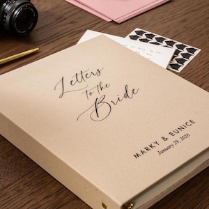 May include: A beige binder with the words "Letters to the Bride" in elegant script. The names "Marky & Eunice" and the date "January 29, 2026" are printed below. A gold pen and camera are visible.