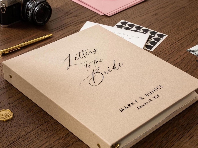 May include: A beige binder with the words "Letters to the Bride" in elegant script. The names "Marky & Eunice" and the date "January 29, 2026" are printed below. A gold pen and camera are visible.