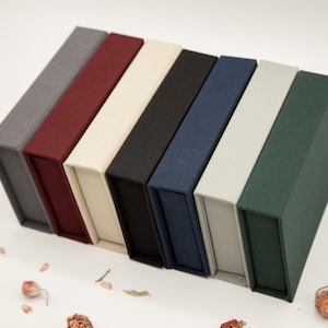 May include: A set of seven small, rectangular boxes in various colors, including gray, burgundy, beige, black, navy blue, light gray, and dark green. The boxes are made of a textured fabric material and are stacked on top of each other.
