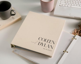 May include: A beige fabric-covered three-ring binder featuring the black printed text "COLEEN and DYLAN" followed by the date "06.15.2023" on the front cover. The binder has gold-toned metal rings and is shown partially open.