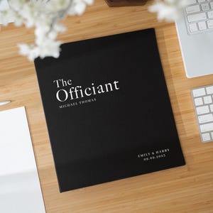 Peut inclure: Un dossier noir avec le texte "The Officiant" en blanc, ainsi que le nom "Michael Thomas". Le bas du dossier affiche "Emily & Harry 09.09.2023". Le dossier est sur une surface en bois, à côté d'un ordinateur portable et d'un clavier.