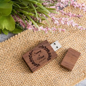 May include: A wooden USB flash drive with a personalized inscription. The inscription reads "Sammy & Mark 01.01.2019" and is surrounded by a wreath of leaves. The drive is sitting on a burlap surface.