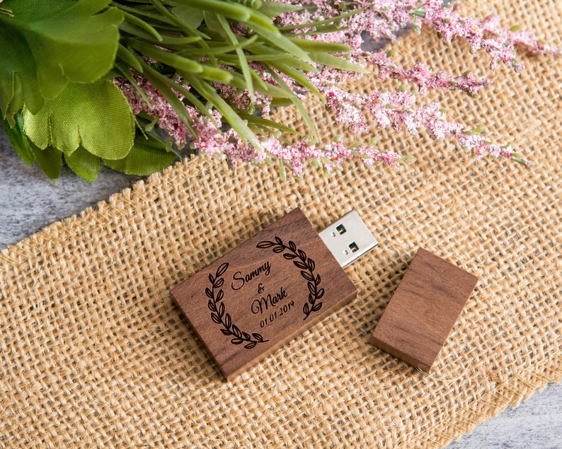 May include: A wooden USB flash drive with a personalized inscription. The inscription reads "Sammy & Mark 01.01.2019" and is surrounded by a wreath of leaves. The drive is sitting on a burlap surface.