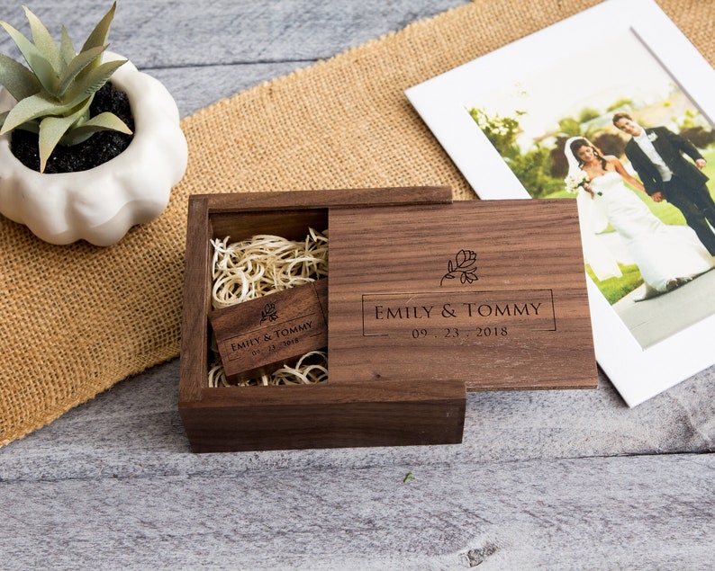 May include: A wooden box with a hinged lid, engraved with the text "Emily & Tommy 09.23.2018" and a floral design. The box contains a wooden USB drive with the same text and design.