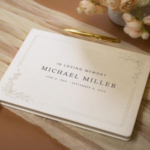 May include: Cream-coloured memorial guest book with the text "IN LOVING MEMORY MICHAEL MILLER JUNE 2, 1950 - SEPTEMBER 8, 2023." Features a decorative floral corner design and a cream-coloured ribbon bookmark. A gold pen rests nearby.