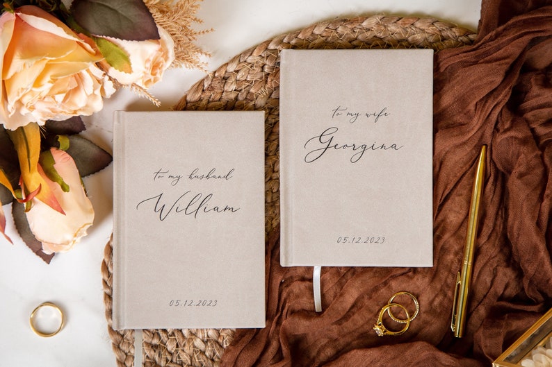 May include: Two beige notebooks with the text "To my husband William" and "To my wife Georgina" in elegant script. The date "05.12.2023" is printed below the names. A gold pen, rings, and flowers are also in the image.