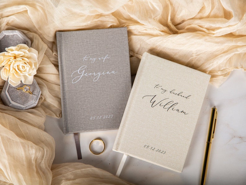 May include: Two hardcover journals with personalized text, one gray and one cream-colored, are displayed on a marble surface. The gray journal reads "To my wife Georgina" and the cream journal reads "To my husband William". A gold pen and wedding ring are also visible.