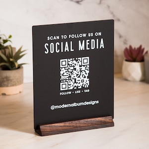 May include: Black sign with white text that reads "SCAN TO FOLLOW US ON SOCIAL MEDIA". A QR code is below the text. Below the QR code is the text "FOLLOW - LIKE - TAG" and "@modernalbumdesigns". The sign is on a wooden stand.