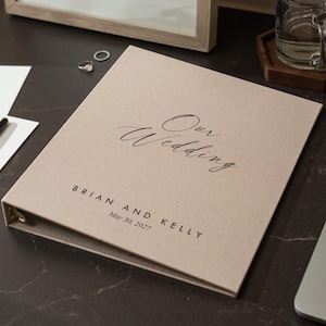 May include: A beige fabric-covered three-ring binder featuring the black printed text "Our Wedding" in a script font, followed by "BRIAN AND KELLY" and the date "May 30, 2027" in a clean, sans-serif typeface centered on the front cover.