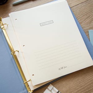 May include: Open light blue binder with gold rings, holding cream-colored pages. The pages have lined spaces and the text "add photo or card here" and "With love." A light blue envelope is visible.