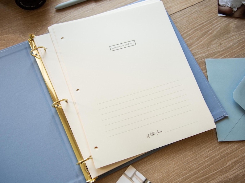 May include: Open light blue binder with gold rings, holding cream-colored pages. The pages have lined spaces and the text "add photo or card here" and "With love." A light blue envelope is visible.