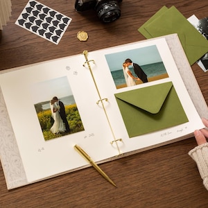 May include: An open scrapbook with wedding photos, green envelopes, and a gold pen on a wooden surface. The scrapbook has a floral design on the cover and gold rings. A camera and stickers are also visible.