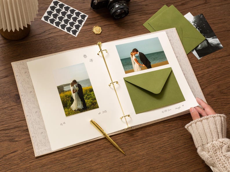 May include: An open scrapbook with wedding photos, green envelopes, and a gold pen on a wooden surface. The scrapbook has a floral design on the cover and gold rings. A camera and stickers are also visible.