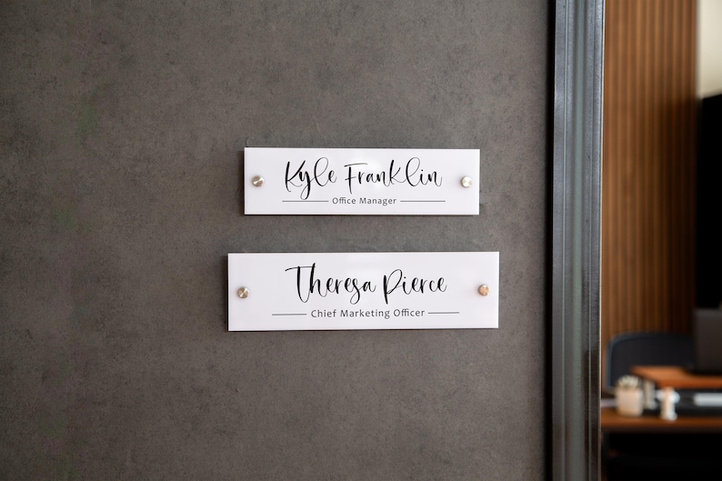 May include: Two white rectangular office door signs with black text. The top sign reads "Kyle Franklin - Office Manager". The bottom sign reads "Theresa Pierce - Chief Marketing Officer".