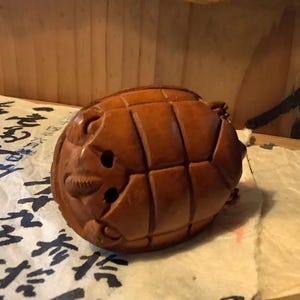 May include: A carved wooden turtle figurine with a detailed shell pattern and facial features. The turtle is a warm brown color and appears to be resting on a piece of paper with Asian characters.