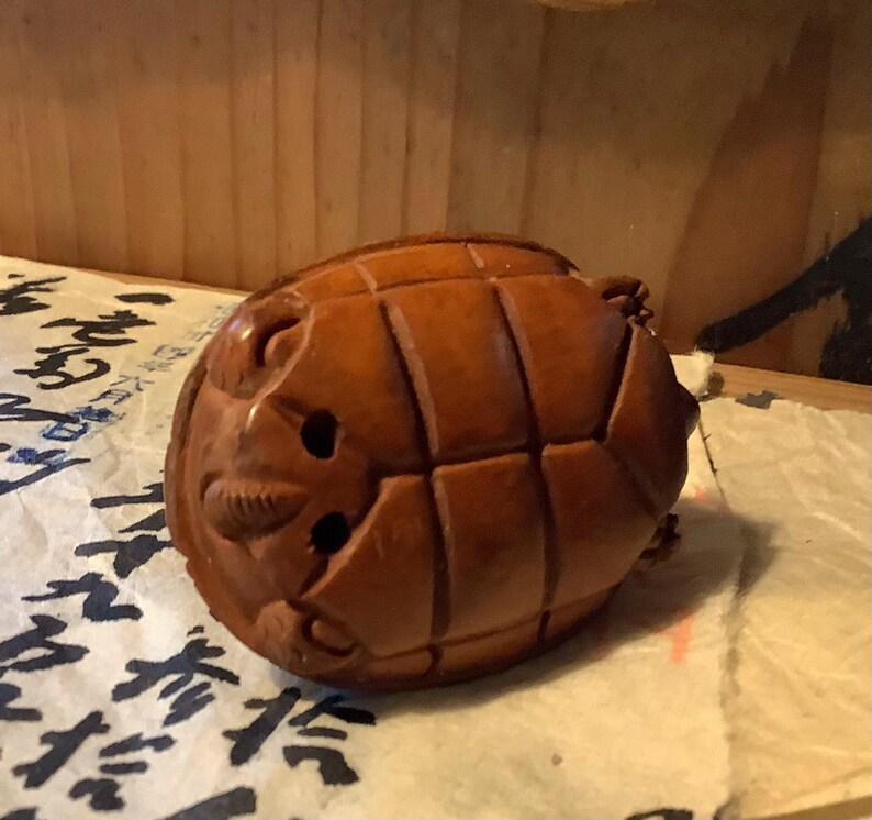 May include: A carved wooden turtle figurine with a detailed shell pattern and facial features. The turtle is a warm brown color and appears to be resting on a piece of paper with Asian characters.