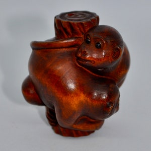 May include: A carved wooden sculpture of a monkey hugging a tree stump. The monkey is brown with black eyes and is embracing a cylindrical tree trunk. The wood has a rich, reddish-brown color and visible grain.