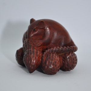 Puede incluir: Una escultura de madera tallada a mano de una rata marrón sentada sobre un montón de nueces. La rata está detallada con un pequeño ojo negro y un patrón de pelaje texturizado.