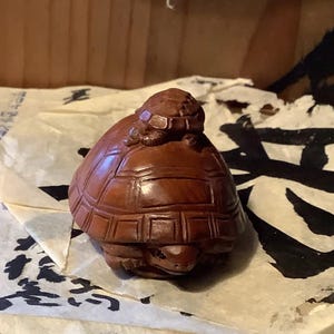 May include: A carved wooden sculpture of a turtle with a smaller turtle on its back. The sculpture is a warm brown color and has detailed carvings of the turtles' shells and faces. The sculpture is resting on a piece of paper with black ink.