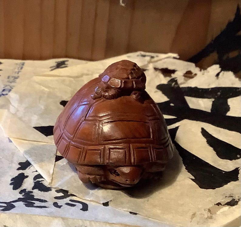 May include: A carved wooden sculpture of a turtle with a smaller turtle on its back. The sculpture is a warm brown color and has detailed carvings of the turtles' shells and faces. The sculpture is resting on a piece of paper with black ink.