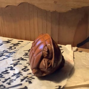 May include: A carved wooden sculpture of a small, rounded object with intricate details. The sculpture is a warm brown color and appears to be resting on a piece of paper with black calligraphy. The wood grain is visible, adding texture to the piece.