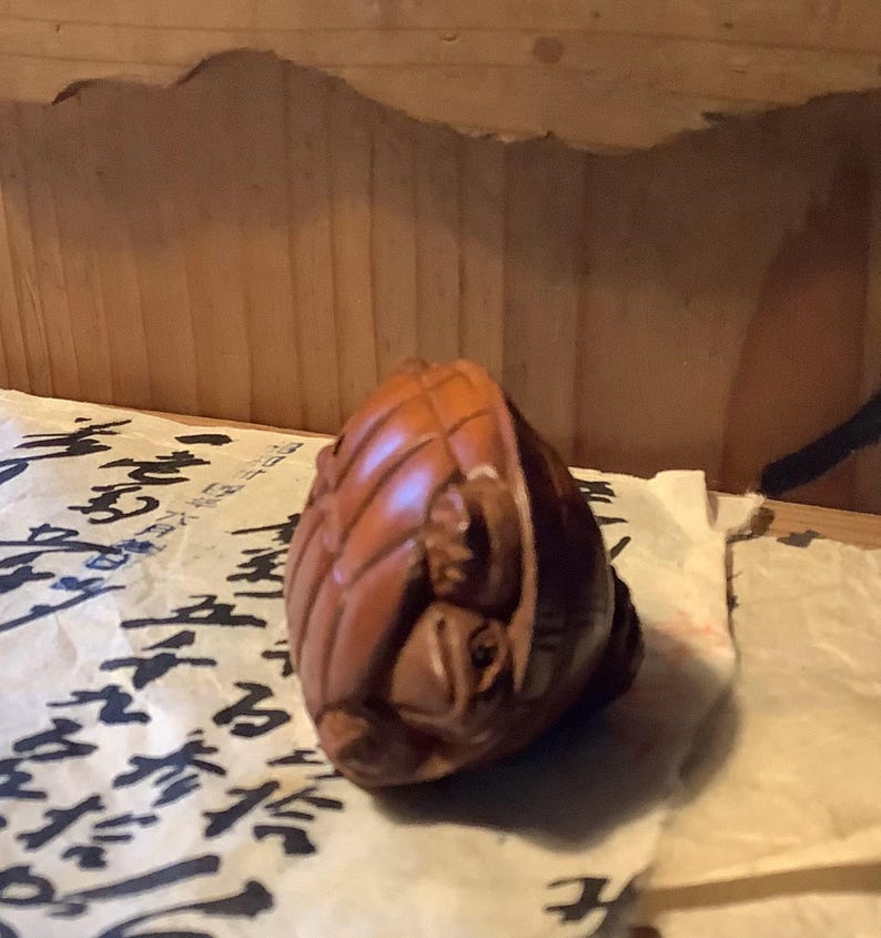 May include: A carved wooden sculpture of a small, rounded object with intricate details. The sculpture is a warm brown color and appears to be resting on a piece of paper with black calligraphy. The wood grain is visible, adding texture to the piece.