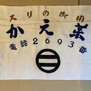 May include: White fabric banner with blue and brown Japanese characters, a black circle with three horizontal lines, and the numbers 2693. The text reads "The Little Shop of the Old Man".