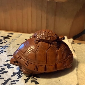 Puede incluir: Una detallada talla de madera de una tortuga grande con una tortuga más pequeña sobre su espalda. Las tortugas son de color marrón cálido, con intrincados patrones de caparazón. La talla está sobre una superficie con escritura asiática.