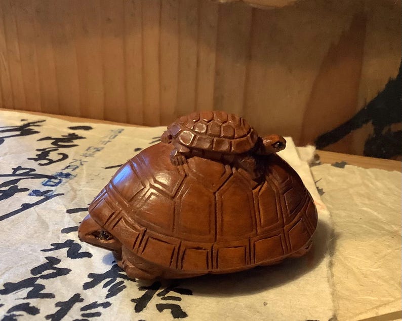 May include: A detailed wooden carving of a large turtle with a smaller turtle on its back. The turtles are a warm brown color, with intricate shell patterns. The carving is set on a surface with Asian script.