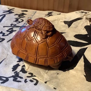 May include: A wooden sculpture of a large turtle with a smaller turtle riding on its back. The turtles are a warm brown color and have detailed shell patterns. The sculpture is set on a piece of paper with black calligraphy.