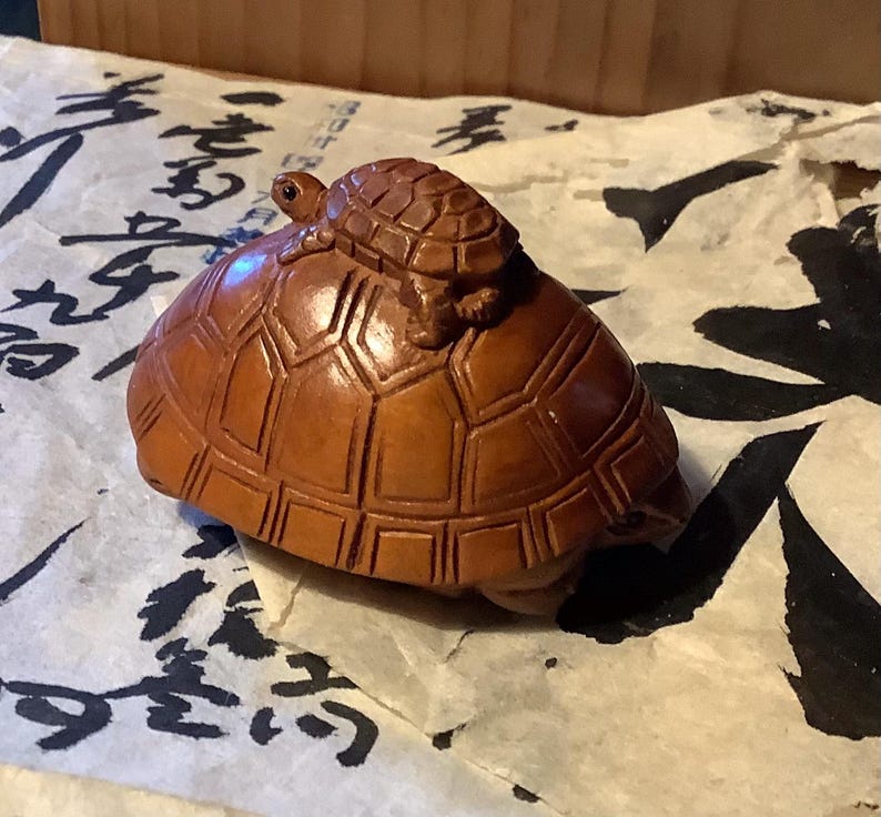 May include: A wooden sculpture of a large turtle with a smaller turtle riding on its back. The turtles are a warm brown color and have detailed shell patterns. The sculpture is set on a piece of paper with black calligraphy.