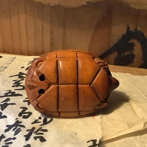 May include: A carved wooden turtle figurine with a detailed shell pattern and a small head. The turtle is a warm brown color and appears to be handmade. The figurine is resting on a piece of paper with black calligraphy.