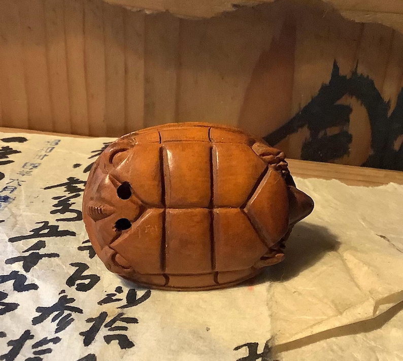 May include: A carved wooden turtle figurine with a detailed shell pattern and a small head. The turtle is a warm brown color and appears to be handmade. The figurine is resting on a piece of paper with black calligraphy.