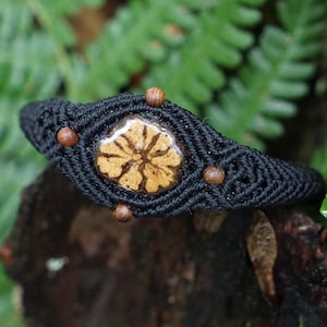 Può includere: Bracciale in macramè nero con un motivo floreale centrale color beige. Il bracciale presenta piccole perline di legno ed è ambientato su uno sfondo di fogliame verde, creando un'estetica naturale.