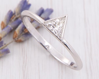 May include: A silver-toned metal ring featuring a single, clear, triangular-cut gemstone set in a bezel setting. The band is smooth and polished, showcasing a clean, geometric design that highlights the central triangular stone.