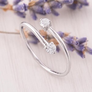 May include: A bypass style ring crafted from polished silver-toned metal, featuring two round, clear, faceted stones set at each open end of the band. The design showcases a sleek, minimalist aesthetic with a continuous, slender, circular metal wire.