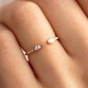 Op de afbeelding: Open gouden ring met twee marquise-geslepen edelstenen. Aan elk uiteinde zit een edelsteen, met een kleine cluster van kleinere stenen aan één kant. De ring wordt aan een vinger gedragen.