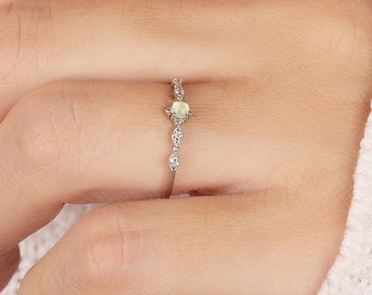 May include: A delicate silver ring with a small, light green opal gemstone set in the center. The band is adorned with tiny, sparkling diamonds.