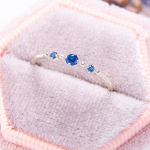 May include: A delicate silver ring with a row of blue sapphires set in a curved band. The ring is displayed in a pink velvet ring box.