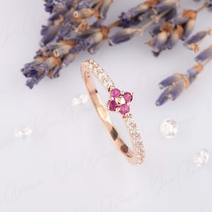 以下が含まれることがあります： 小さな花のデザインが施された繊細なローズゴールドのリング。ピンクの宝石が5つ、小さな白い宝石で囲まれています。